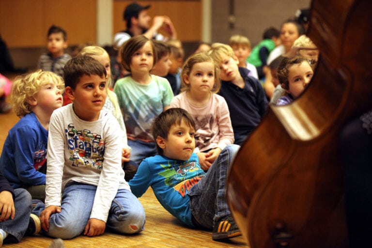 Einige Kinder sitzen auf dem Boden während sie einer Person mit Cello zuhören