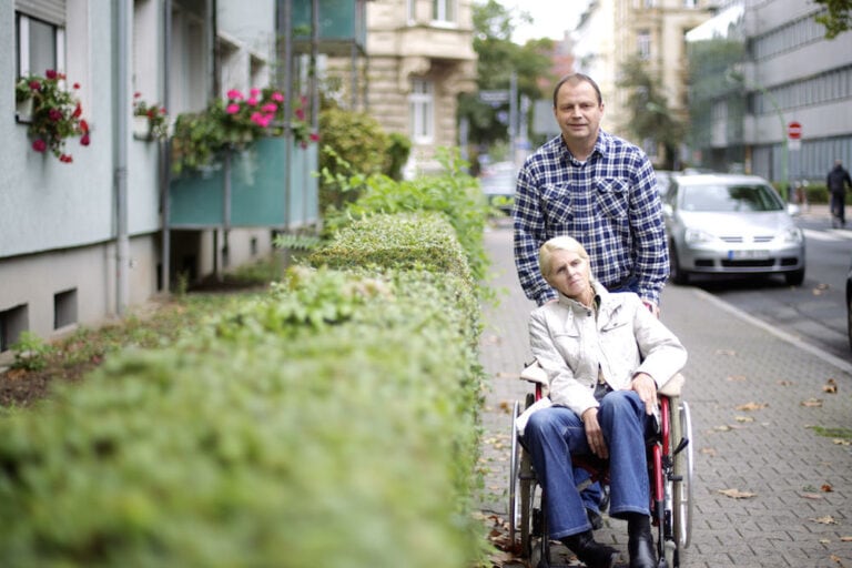 Mann schiebt ältere Frau im Rollstuhl auf einem Gehweg