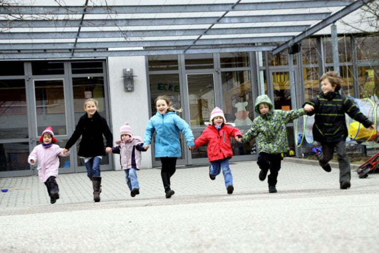 Sieben Kinder in herbstlicher Kleidung rennen Hand in Hand auf dem Fotografen zu.