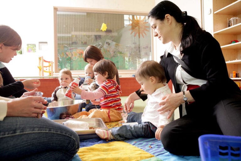 Mehrere Erzieherinnen sitzen mit Kindern auf einer Decke auf dem Boden
