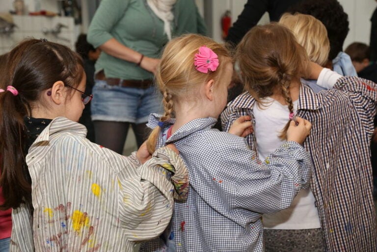 Vier Kinder stehen hintereinander und machen dem jeweils vorderen Kind das falsch herum getragene Malerhemd zu.