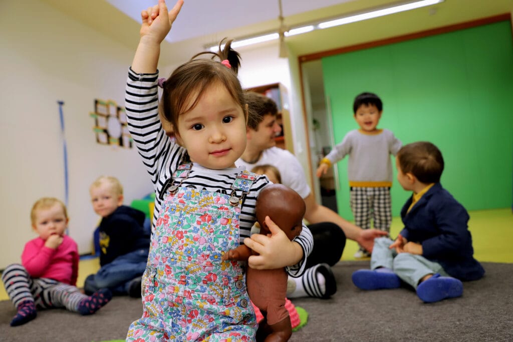 Ein Mädchen mit einer Puppe kniet in einem Kita-Raum mit anderen Kindern und streckt den Finger.