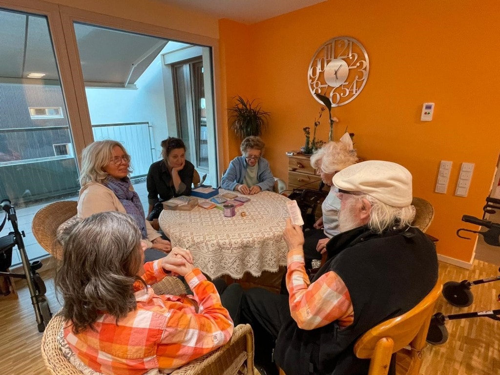 Ältere Menschen sitzen an einem runden Tisch in einem Raum mit orangenen Wänden.