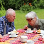 Ein älterer Mann und eine ältere Frau sitzen an einem Gartentisch, trinken Kaffee und essen Kuchen.
