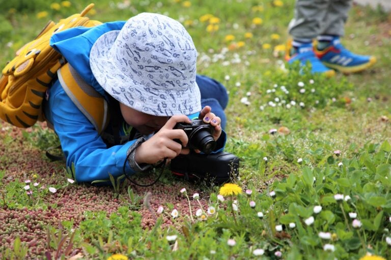 Ein Kind kniet tief auf dem Boden und fotografiert eine Blume.