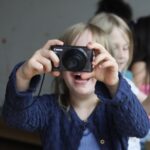 Ein blondes Kind hält seine Kamera vor sein Gesicht und schießt lachend ein Foto.