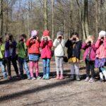 Acht Mädchen und ein Junge stehen nebeneinander auf einem Waldweg und halten alle ihre Kameras vor ihre Gesichter.
