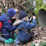 Ein Junge und ein Mädchen knien vor einem großen Betonrohr im Wald und schießen mit ihren Kameras Fotos.
