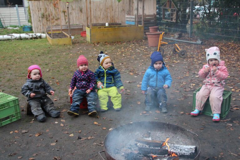 fünf Kinder in Matschhosen, Jacken und Mützen sitzen vor einem Lagerfeuer